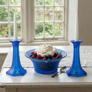 May include: Two blue glass candle holders flank a bowl of berries topped with whipped cream. A silver spoon rests near the bowl. The candle holders have a hexagonal base and floral accents. A window is in the background.