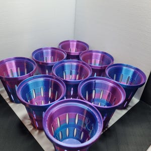Op de afbeelding: Een verzameling van tien kleine, cilindervormige plantenpotten. De potten hebben een kleurovergang van paars, blauw en roze. Elke pot heeft een reeks verticale, ovale gaten. De potten zijn in een groep gerangschikt, sommige van bovenaf en andere van de zijkant zichtbaar.