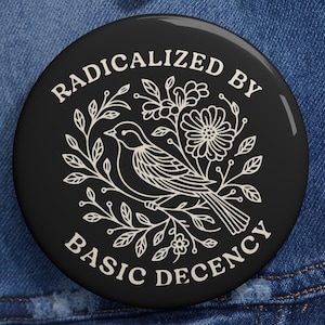 Może przedstawiać: Czarny guzik z białym napisem "RADICALIZED BY BASIC DECENCY", otaczającym białą ilustrację ptaka i kwiatów. Guzik jest umieszczony na niebieskiej kurtce dżinsowej.