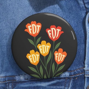 Puede incluir: Un botón redondo negro con un diseño floral. El diseño presenta cinco flores estilizadas de color naranja y amarillo con las letras "FDT" en blanco. El botón está sobre un fondo de mezclilla.
