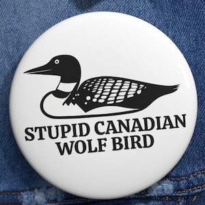 May include: A white button with a black and white illustration of a loon and the text "STUPID CANADIAN WOLF BIRD". The button is round and has a glossy finish. The design is simple and bold, with a humorous tone.