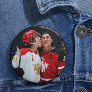May include: A round button featuring two hockey players in red and white jerseys. One player is kissing the other on the cheek. The button is set against a blue denim jacket.