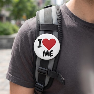 Puede incluir: Una chapa redonda de color blanco con el texto en negro I y ME y un gran símbolo de corazón rojo en el centro. La chapa está sujeta a la correa gris y negra de una mochila, mostrando claramente la frase I corazón ME en la superficie.