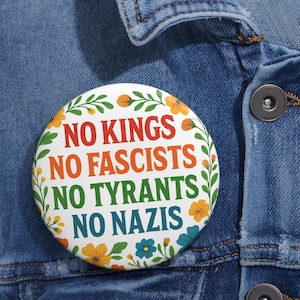 Puede incluir: Un botón redondo blanco con el texto "NO KINGS NO FASCISTS NO TYRANTS NO NAZIS" en rojo, naranja, verde y azul. El botón está rodeado por un diseño floral con flores naranjas y amarillas y hojas verdes. El botón está prendido a una chaqueta vaquera.