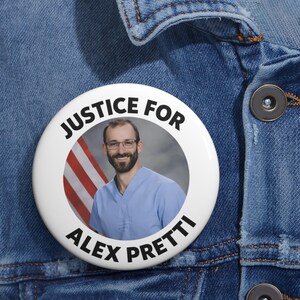 May include: A white button with the text "JUSTICE FOR ALEX PRETTI" in black, encircling a photo of a man in glasses and a blue shirt, with an American flag in the background. The button is pinned to a blue denim jacket.