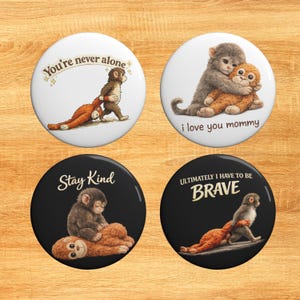 May include: Four circular badges featuring monkey designs. Two are white, with the phrases "You're never alone" and "i love you mommy". The other two are black, with the phrases "Stay Kind" and "ULTIMATELY I HAVE TO BE BRAVE".