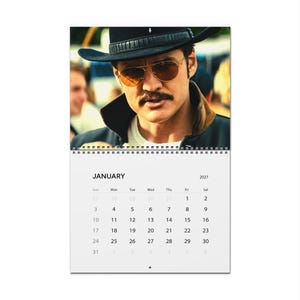 May include: A wall calendar for January 2027 featuring a photograph of a person wearing a black cowboy hat and gold-tinted aviator sunglasses. The calendar page displays the month name, year, and a grid of dates from the first to the thirty-first.