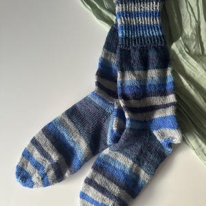 Könnte beinhalten: Ein Paar handgestrickte Socken in Blau- und Grautönen. Die Socken haben horizontale Streifen und ein geripptes Bündchen. Das Garn scheint eine Mischung aus Wolle oder einer ähnlichen Faser zu sein. Die Socken werden auf einer weißen Oberfläche mit einem grünen Stoff im Hintergrund präsentiert.