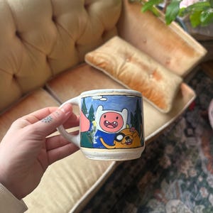 May include: A white ceramic mug with a black handle and the text "But First, Coffee" in a bold, black font. The mug is set against a blurred background, suggesting a cozy setting. The mug appears to be approximately 4 inches tall.