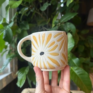 Puede incluir: Taza de cerámica color crema con un diseño de margarita amarilla. La taza tiene un centro negro y un asa curva. El diseño de la margarita está en relieve en la superficie de la taza. La taza está sostenida en la mano de una persona.