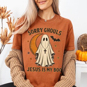 May include: A burnt orange short-sleeve t-shirt featuring a graphic of a ghost wearing a cross necklace, a crescent moon, a bat, and stars. The text reads SORRY GHOULS JESUS IS MY BOO in black lettering. The shirt is worn under a tan knit cardigan.