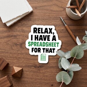 May include: A sticker with the text "RELAX, I HAVE A SPREADSHEET FOR THAT" in black and green. The sticker has a white border and a spreadsheet icon. The sticker is on a wooden surface with other office supplies.