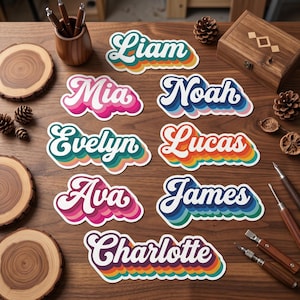 May include: A collection of retro-style name stickers on a wooden surface. Each sticker features a different name, including "Liam," "Mia," and "Noah," in a white, bold font with colorful, layered outlines. The stickers are arranged with other craft supplies.