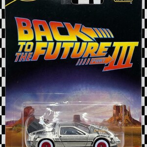 Pode incluir: Um carro de brinquedo Hot Wheels Pop Culture com o DeLorean de Back to the Future Part III. O carro prateado tem rodas vermelhas e é exibido em um cartão com o logotipo do filme e um cenário desértico.
