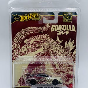 Peut inclure: Une voiture miniature Hot Wheels Premium Nissan Skyline GT-R (BNR34) dans un étui en plastique transparent. L'emballage présente un thème Godzilla avec des couleurs rouge et beige et le texte "GODZILLA" et "POP CULTURE".