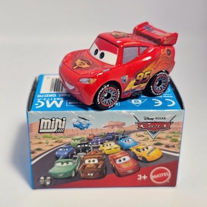 Może przedstawiać: Czerwony samochodzik Lightning McQueen z numerem 95 z boku, umieszczony na pudełku z r&oacute;żnymi postaciami z filmu Auta. Pudełko jest oznaczone jako "mini" i "Disney Pixar Cars". Samochodzik ma czarno-srebrne koła.