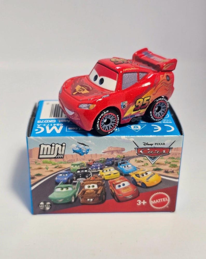 Może przedstawiać: Czerwony samochodzik Lightning McQueen z numerem 95 z boku, umieszczony na pudełku z r&oacute;żnymi postaciami z filmu Auta. Pudełko jest oznaczone jako "mini" i "Disney Pixar Cars". Samochodzik ma czarno-srebrne koła.