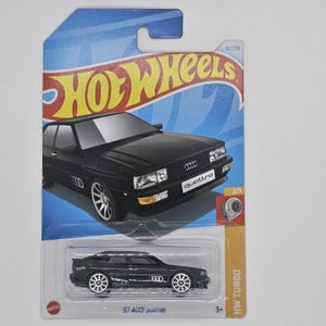 以下が含まれることがあります： ホットウィールカード上の黒い'87 Audi Quattroダイキャストモデルカー。車は白いホイールで、前面に「Quattro」の文字があります。上部にはHot Wheelsのロゴが見えます。