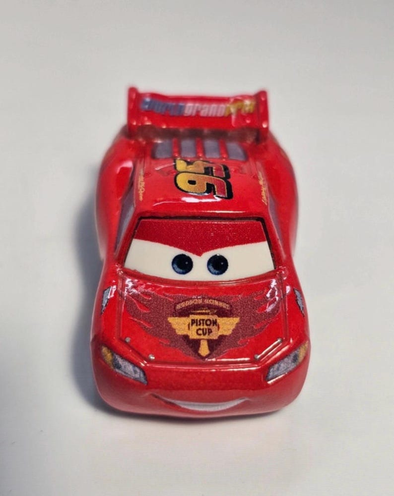 Może przedstawiać: Czerwony samochodzik z numerem 95 w kolorze złotym oraz napisami "Rust-eze" i "Piston Cup". Samoch&oacute;d ma niebieskie oczy i spojler. Zabawkowy samoch&oacute;d to replika Zygzaka McQueena z filmu Auta.