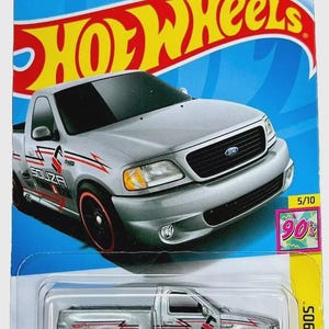 以下が含まれることがあります： シルバーのホットウィールのおもちゃの車、'99 Ford F-150 SVT Lightningピックアップトラック。トラックは赤と黒のアクセント、赤いリムの黒いホイール、そして「SOUZA Plumbing & Heating」の文字があります。パッケージは青と黄色です。