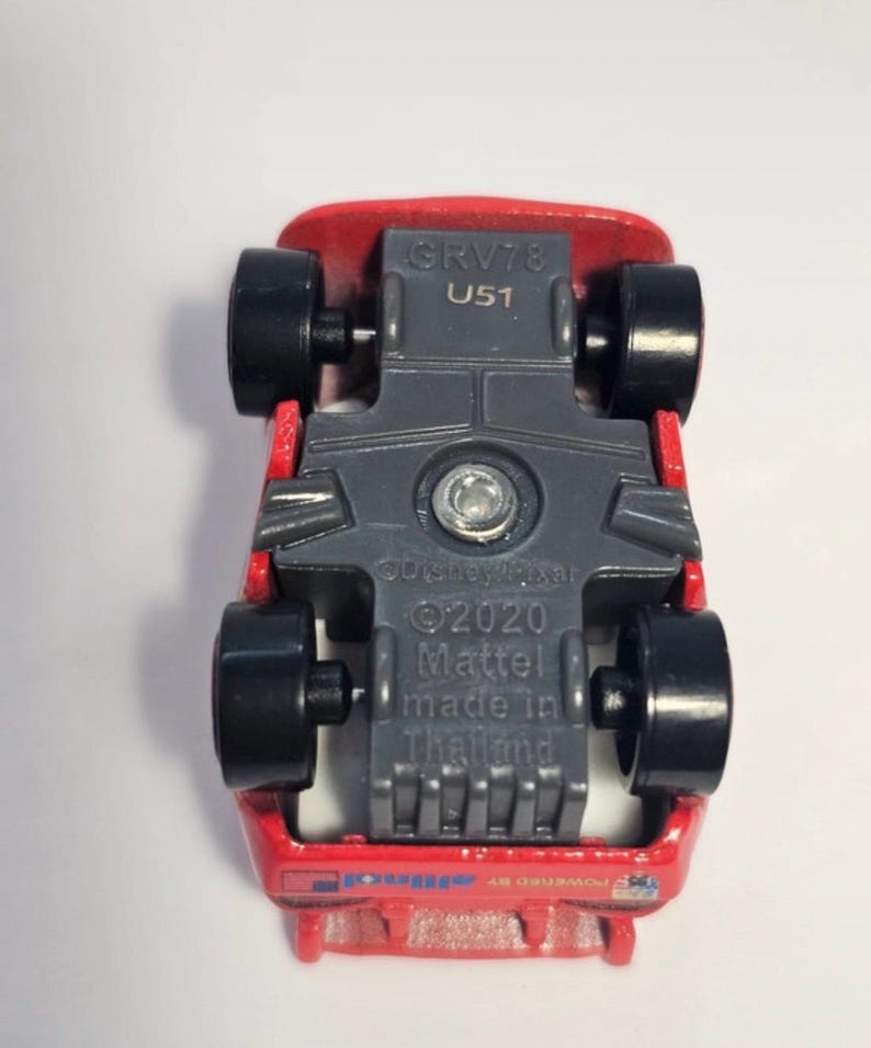 Może przedstawiać: Czerwony odlewany samoch&oacute;d zabawkowy z czarnymi kołami, widok od spodu. Samoch&oacute;d ma szare podwozie z napisem "GRV78 U51", "&copy;Disney/Pixar &copy;2020 Mattel made in Thailand" i "powered by allinol".