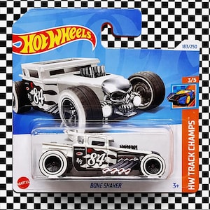 以下が含まれることがあります： 青と白のパッケージに入った、Hot Wheels Bone Shakerのダイキャストカー。車体は白で黒のアクセント、84の数字、スカル型のフロントが特徴です。パッケージにはHot Wheelsのロゴと「HW Track Champs」が含まれています。