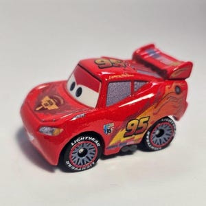 Może przedstawiać: Czerwony samochodzik Lightning McQueen z numerem 95 na dachu i bokach. Samoch&oacute;d ma spojler, a na oponach widnieje napis Lightning McQueen. Samoch&oacute;d ma ż&oacute;łto-pomarańczowy płomień po boku.