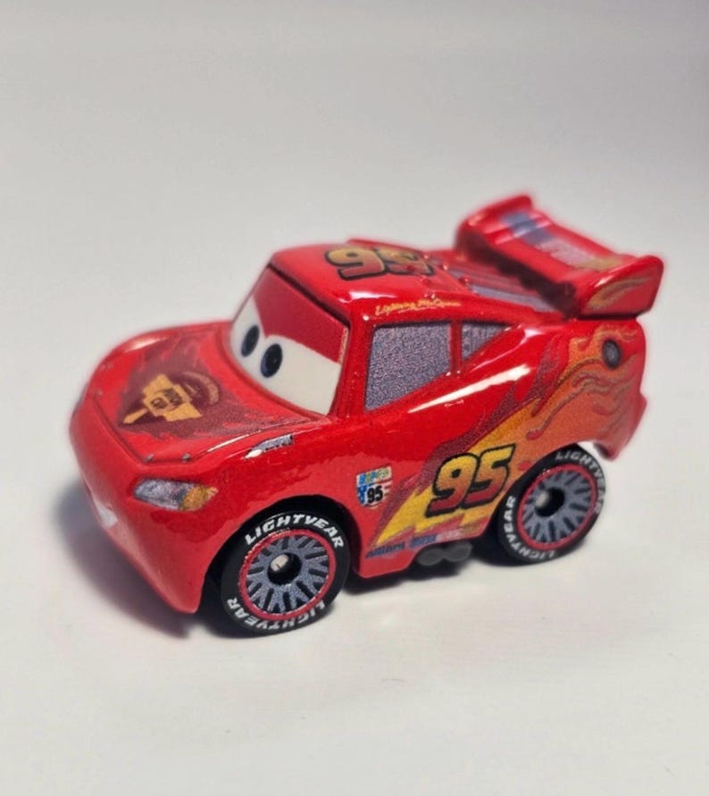 Może przedstawiać: Czerwony samochodzik Lightning McQueen z numerem 95 na dachu i bokach. Samoch&oacute;d ma spojler, a na oponach widnieje napis Lightning McQueen. Samoch&oacute;d ma ż&oacute;łto-pomarańczowy płomień po boku.