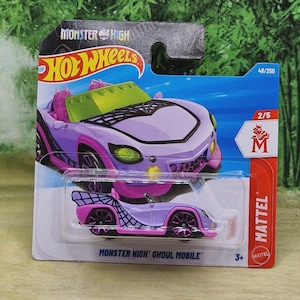 以下が含まれることがあります： パッケージに入ったHot Wheelsのモンスターハイ・グールモバイルのおもちゃの車。車体は紫とピンクで、クモの巣のデザイン。パッケージにはHot Wheelsのロゴと「Monster High Ghoul Mobile」の文字が入っています。