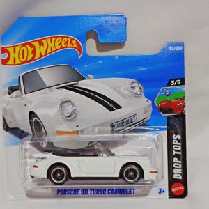 以下が含まれることがあります： 黒いレーシングストライプと黒いホイールが付いた白いポルシェ911ターボカブリオレのおもちゃの車。おもちゃの車は、青と白のホットウィールパッケージに入っています。「PORSCHE 911 TURBO CABRIOLET」という文字がパッケージに印刷されています。