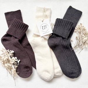 May include: Three pairs of ribbed knit socks in dark brown, cream, and charcoal gray. The cream pair features a white square tag with the text EN Elegant Neutrals 100% Merino Wool. Each pair has a folded cuff and a textured, vertical ribbed pattern.