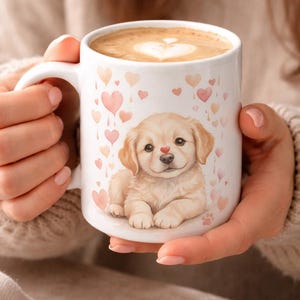 Puede incluir: Taza de cerámica blanca con una ilustración de cachorro golden retriever, una nariz de corazón rojo y corazones rosas. La taza contiene una bebida de café con un diseño en forma de corazón en la espuma. Una huella de pata está en la parte inferior de la taza.
