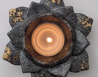 Puede incluir: Un portavelas negro de cerámica con forma de flor de loto con detalles dorados y una vela encendida en el centro. Los pétalos están salpicados de blanco y dorado, creando un diseño decorativo y elegante.
