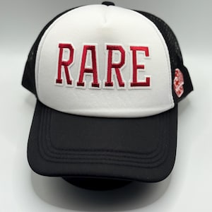 Könnte beinhalten: Schwarz-weiße Trucker-Cap mit dem Wort "RARE" in roten Großbuchstaben auf der Vorderseite gestickt. Die Kappe hat einen schwarzen Schirm und eine Netzrückseite mit einer weißen Frontplatte. Ein kleines rotes Emblem befindet sich an der Seite.