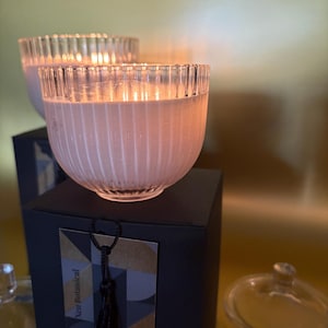 Puede incluir: Dos velas encendidas en cuencos de vidrio transparente y acanalado, apoyadas sobre cajas negras. Las velas tienen un suave tono rosado, y las cajas tienen una etiqueta con el texto "Xen Botanical" y una borla negra. También se ve una tapa de cristal transparente.