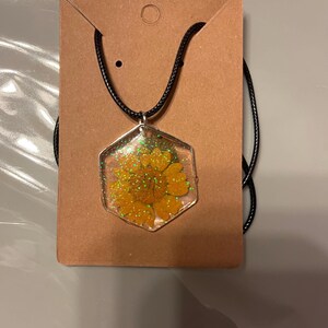 Puede incluir: Un collar con colgante hexagonal que contiene una flor amarilla en resina transparente y purpurina dorada. El colgante cuelga de un cordón negro y se muestra en una tarjeta marrón.