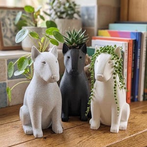 以下が含まれることがあります： 座った姿勢の犬の形をしたプランター3つ。左のプランターは質感のあるライトグレー、中央は黒、右は白です。それぞれのプランターの上部には空洞があり、小さな緑の植物が植えられています。
