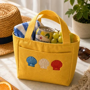 May include: A yellow tote bag with embroidered seashell designs in blue, white, and red. The bag contains a striped towel, a can of Sanpellegrino, and grapes. A straw hat and sunglasses are in the background.