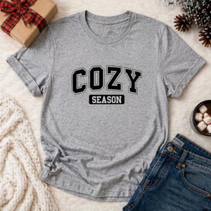 May include: A heather gray short-sleeve t-shirt featuring the text COZY SEASON in black block lettering across the chest. The shirt has a crew neckline and a standard fit, presented as a casual garment with a simple, clean design.