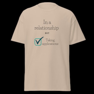 May include: Beige t-shirt with the text "In a relationship BUT Taking applications". The text is in black and teal. The shirt has a crew neck and short sleeves.