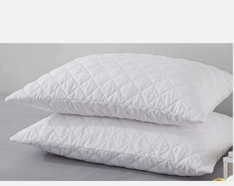 May include: Two white quilted pillows stacked on top of each other. The pillows have a diamond pattern and are made of a soft, smooth material. The pillows are on a light gray surface.