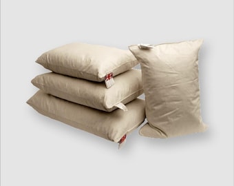 May include: Four beige pillows stacked and a fifth pillow standing upright. Each pillow has a tag with a red and white design. The pillows are a neutral color and appear to be made of a soft material.