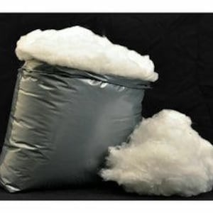 May include: A silver-grey bag filled with fluffy, white polyester fibre, with a smaller pile of the same material beside it. The bag is open at the top, revealing the contents. The background is a solid black.