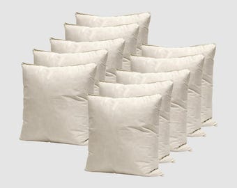 May include: A collection of ten square, off-white pillows arranged in a layered display. The pillows have a soft, textured appearance and a neutral color palette, suitable for various home decor styles. The seams are visible.