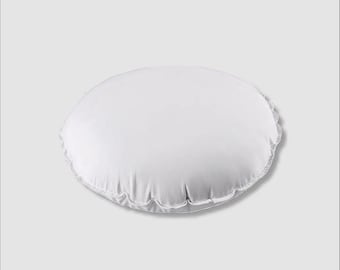 May include: A white, round pillow with a ruffled edge. The pillow is made of a soft, smooth fabric and appears to be filled with a fluffy material. The pillow is a solid white color and is set against a light gray background.