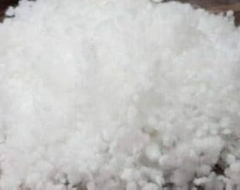 May include: A large pile of fluffy, white polyester fiberfill. The material has a soft, cloud-like appearance and is likely used for stuffing pillows, cushions, or other craft projects. The fiberfill is shown on a wooden surface.