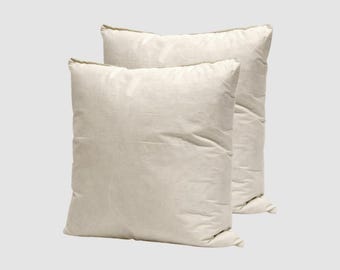 May include: Two off-white square pillows. The pillows are made of a soft, textured fabric and have a slightly rumpled appearance. The pillows are positioned against a light gray background, with one pillow slightly behind the other.