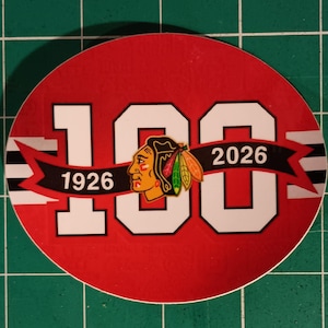 Puede incluir: Una pegatina redonda con fondo rojo y el número "100" en blanco. La pegatina presenta una pancarta negra con los años "1926" y "2026" y un logotipo de una cabeza nativa americana. La pegatina tiene un borde blanco.