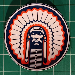 Op de afbeelding: Een ronde sticker met een afbeelding van een Indiaanse stamhoofd met een hoofdtooi. Het ontwerp bevat een blauwe achtergrond, witte veren en oranje accenten. Het gezicht van de stamhoofd is gedetailleerd in donkerblauw en wit.