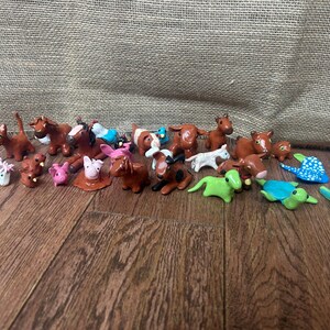 May include: A collection of small, handcrafted animal figurines. The assortment includes horses, pigs, dogs, turtles, and other creatures, all in various colors and poses. The figurines are arranged on a wooden surface.